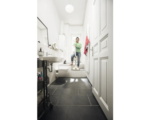 Baie luminoasă cu o femeie sprijinindu-se de un mop fără fir, chiuvetă, toaletă și gresie închisă la culoare.