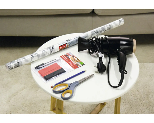 Folie decorativă, racletă, cutter, uscător de păr, creion și foarfece pe o masă