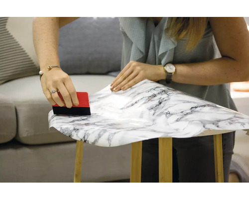 Femeie aplică folie autoadezivă cu aspect de marmură pe un taburet din lemn cu o racletă