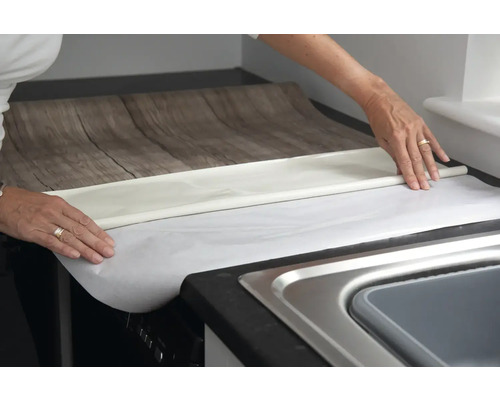 O persoană aplică folie pentru mobilă cu aspect de lemn pe un blat de bucătărie.