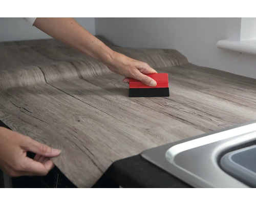 O persoană aplică folie decorativă autoadezivă cu aspect de lemn pe un blat de lucru, folosind o racletă.