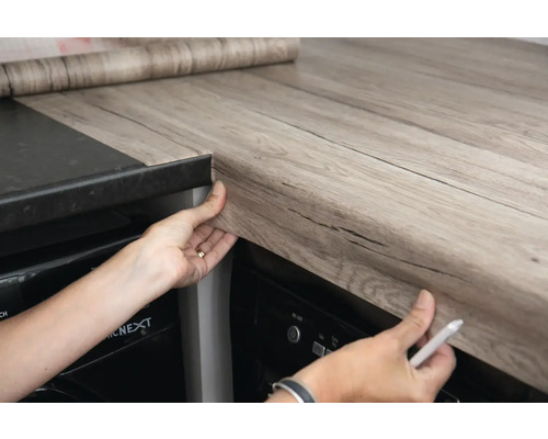 O persoană aplică folie autoadezivă cu aspect de lemn pe un blat de bucătărie.