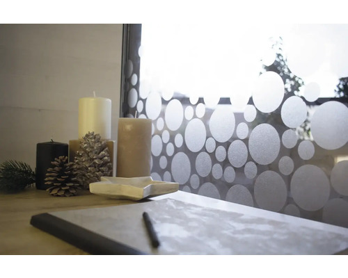 Aranjament decorativ cu lumânări, conuri de pin, bol în formă de stea și instrumente de scris în fața unei ferestre cu model punctat.