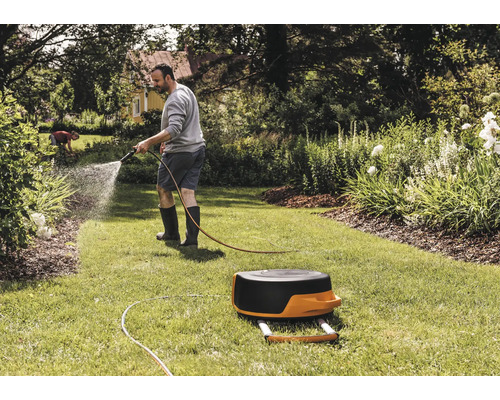 Un bărbat udă cu un furtun de grădină un gazon lângă o rolă de furtun.