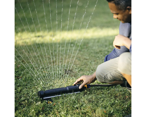 Un bărbat operează un aspersor de gazon în grădină, care pulverizează apă.