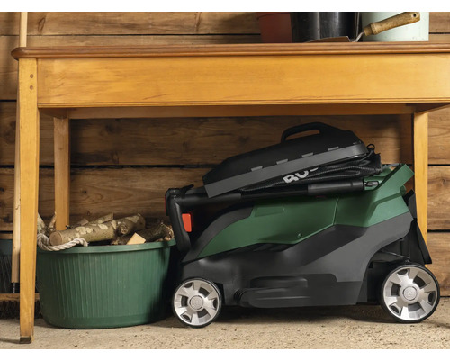 Mașină de tuns iarba pliată sub o masă de lemn într-un garaj.