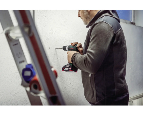 Un bărbat găurește un perete cu o bormașină cu acumulator, lângă o scară.