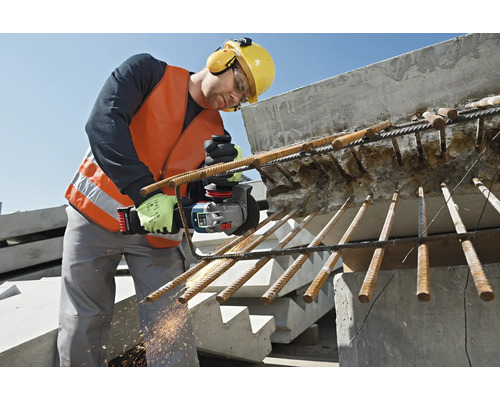 Un muncitor în construcții, cu vestă de protecție, ochelari de protecție, protecție auditivă și cască, taie oțelul de armare cu un polizor unghiular.