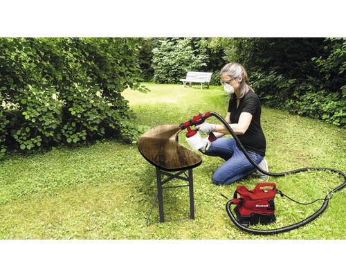 Femeia folosește un sistem de pulverizare a vopselei Einhell pentru a lăcui o bancă de lemn în grădină.