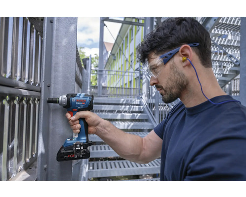 Meșter cu ochelari de protecție și protecție auditivă lucrând cu o șurubelniță cu impact cu acumulator pe o construcție metalică.