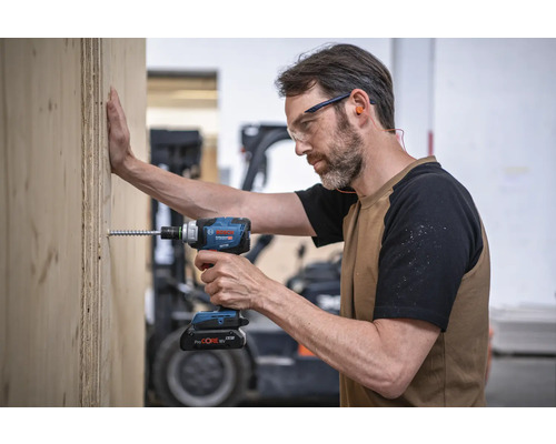 Un bărbat înșurubează un șurub într-o scândură de lemn cu o șurubelniță cu impact cu acumulator. Poartă ochelari de protecție și dopuri de urechi. Logo Bosch.