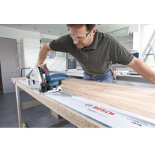 Bărbat taie o scândură de lemn cu un ferăstrău vertical Bosch pe o șină de ghidare.