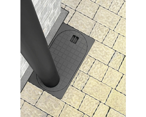 Vedere de sus a unei rigole de curte din plastic cu grătar lângă o conductă de scurgere pe o suprafață pavată.