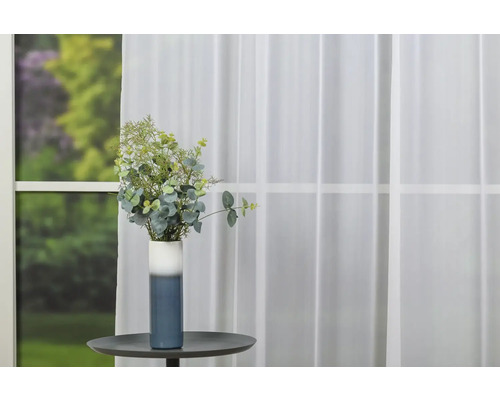 Vază decorativă cu aranjament floral pe o masă în fața unei ferestre cu perdea.