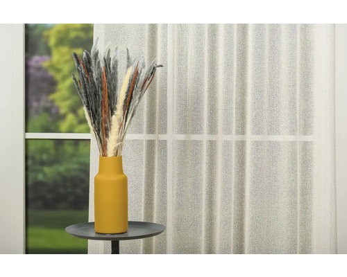 Ierburi decorative într-o vază pe o masă în fața unei ferestre cu perdele