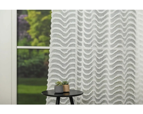 Cortină decorativă albă cu model ondulat în fața unei ferestre, cu o masă și două plante mici.