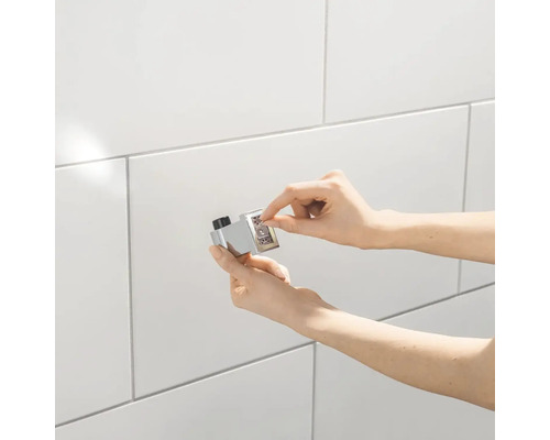 Prim-plan cu o mână care operează un buton termostatic cromat cu numere pe un perete cu gresie