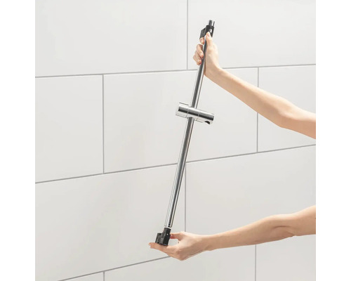 O persoană ține o bară de duș cu suport pentru capul de duș în fața unui perete placat cu faianță.