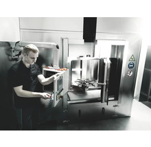 Un bărbat operează un sistem industrial de curățare pentru piese metalice. Semnele de siguranță avertizează asupra pericolelor.