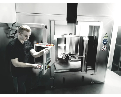 Un bărbat operează un sistem industrial de curățare pentru piese metalice. Semnele de siguranță avertizează asupra pericolelor.