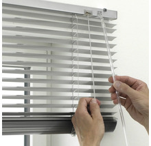 Vedere de aproape a unei mâini care operează o jaluzea din aluminiu argintiu cu o tijă de orientare transparentă și șnur de tragere.