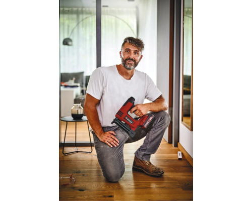 Meșter îngenuncheat pe o podea de lemn, ținând un capsator cu acumulator Einhell, într-un interior modern.