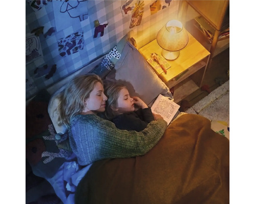 Mama și copilul stau în pat și citesc la lumina difuză a unei veioze într-o cameră de copii confortabilă.