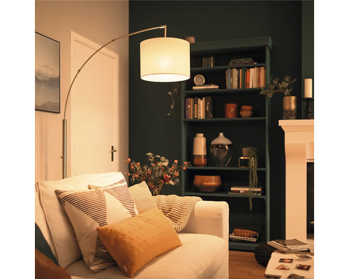 Lampadar arcuit modern cu abajur din material textil lângă o canapea deschisă la culoare într un living confortabil cu bibliotecă închisă.