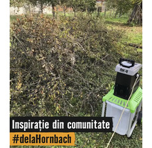 Un tocător de grădină electric marca for_q stă lângă o grămadă mare de ramuri și resturi vegetale pe o peluză.