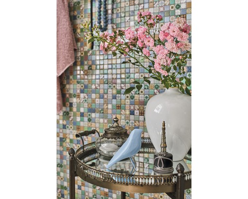 Aranjament decorativ cu flori, pasăre, ceainic și flacon pe o măsuță laterală în fața unui perete de mozaic