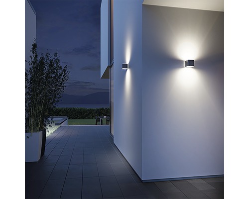 Două lămpi de perete pătrate de pe peretele unei case iluminează o terasă pe timp de noapte.
