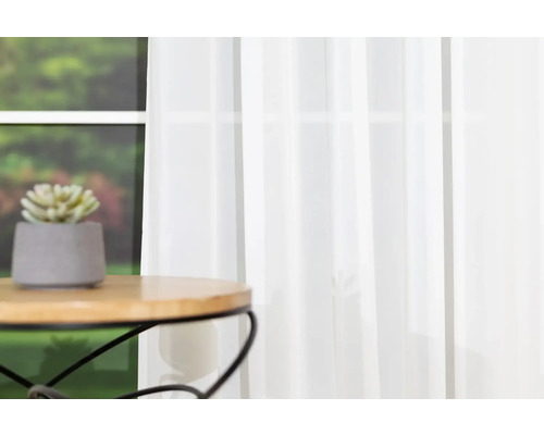 Aranjament decorativ cu plantă în ghiveci pe masă în fața ferestrei cu perdea albă transparentă.