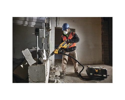 Un bărbat în echipament de protecție folosește un ciocan demolator pe un perete. Lângă el se află un aspirator.