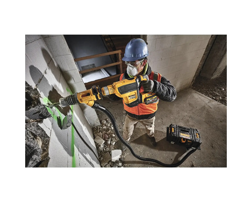 Un muncitor în construcții, în haine de lucru, folosește un ciocan demolator DeWalt pentru a lucra la un perete. Se vede o cutie cu scule.