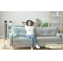 Un ventilator tip turn stă într-un living luminos, lângă o canapea pe care stă o femeie relaxată.