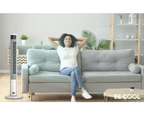 Un ventilator tip turn stă într-un living luminos, lângă o canapea pe care stă o femeie relaxată.
