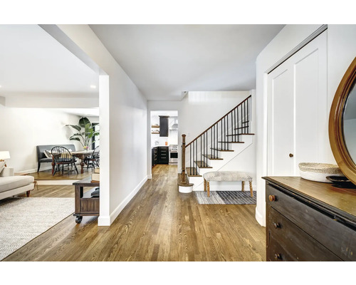 Fotografie de interior a unui hol luminos cu scară, comodă și vedere spre bucătărie și zona de living.