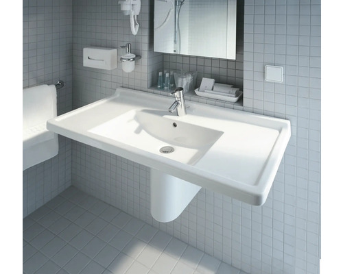 Chiuvetă albă cu robinet în baie, în fața unui perete cu gresie.