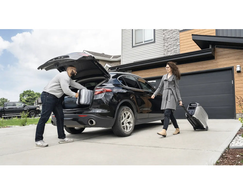 Un bărbat pune valize în portbagajul unei mașini, în timp ce o femeie trage un generator electric portabil pe roți.