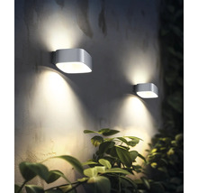 Două lămpi de perete moderne de exterior iluminează un perete lângă o plantă.