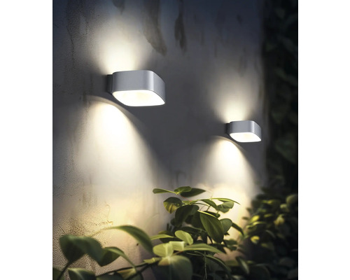 Două lămpi de perete moderne de exterior iluminează un perete lângă o plantă.