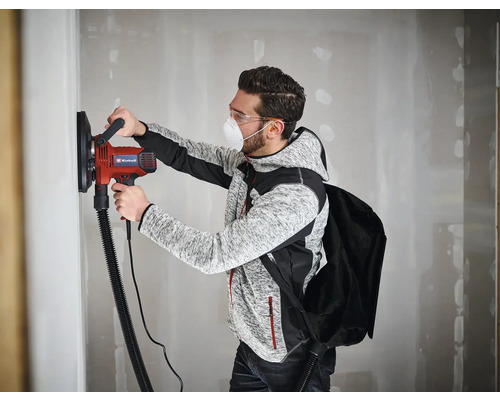 Un bărbat șlefuiește un perete cu un șlefuitor de gips-carton Einhell, poartă mască de praf și ochelari de protecție.