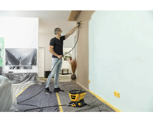 Bărbat care vopsește un perete cu un sistem de pulverizare a vopselei într-o sufragerie protejată cu materiale de acoperire.