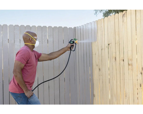 O persoană vopsește un gard din lemn cu un pistol de pulverizare a vopselei, purtând o mască de protecție și ochelari de protecție.