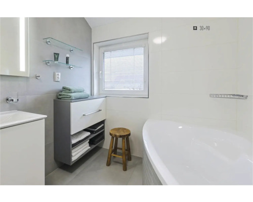 Baie luminoasă cu cadă, lavoar, dulap inferior, prosoape, taburet din lemn și fereastră cu jaluzea