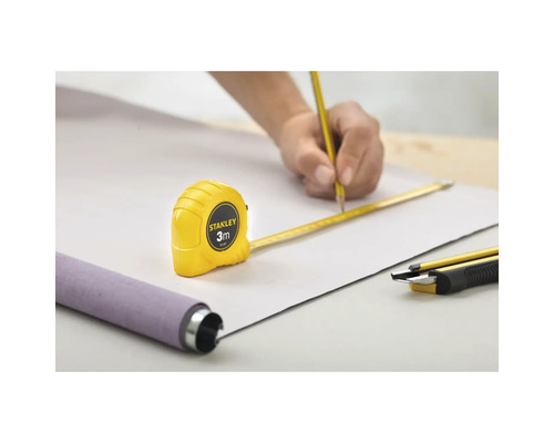Scenă cu o persoană care trasează o linie cu creionul pe o piesă de lucru, asistată de o ruletă Stanley de 3 metri, cu un produs rulat și un cutter în apropiere.