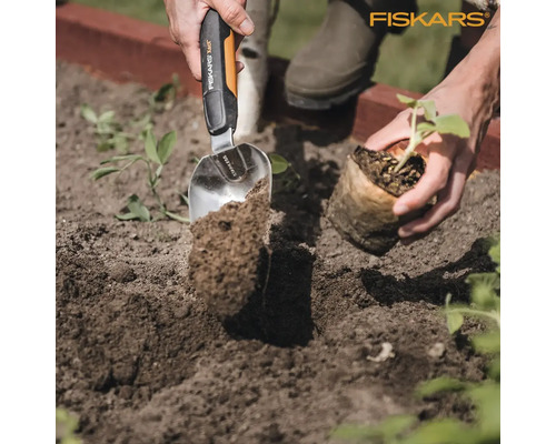 Scenă de grădinărit cu o persoană care plantează o plantă în pământ folosind o lopată din oțel inoxidabil marca Fiskars