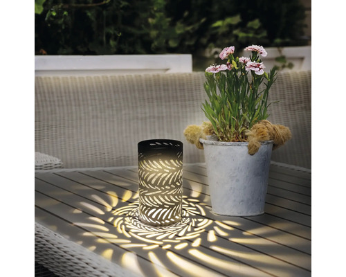 Lampă de masă decorativă cu motiv de frunze lângă un ghiveci cu flori pe o masă de grădină