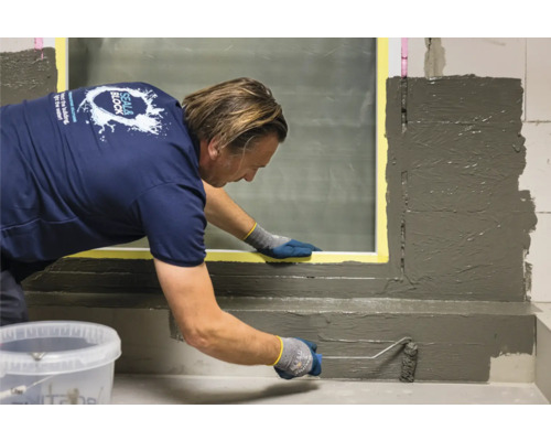 O persoană aplică etanșant cu o rolă de vopsea pe zidărie lângă o fereastră.