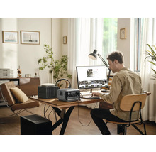 Un bărbat lucrează la un computer cu o centrală electrică portabilă, un router și un dispozitiv de stocare în rețea pe o masă într-o cameră luminoasă.